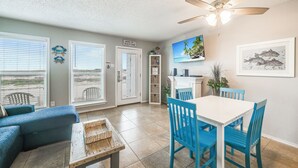 A spacious living area with a white ceiling fan, a dining table with blue chairs, and a view through large windows. - Beachside Retreat | Pool, Hot Tub & Beach Access (Corpus Christi)