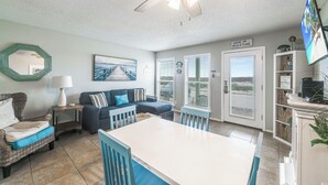 A cozy living room with a dining table, blue chairs, a wicker sofa, and a ceiling fan. - Beachside Retreat | Pool, Hot Tub & Beach Access (Corpus Christi)