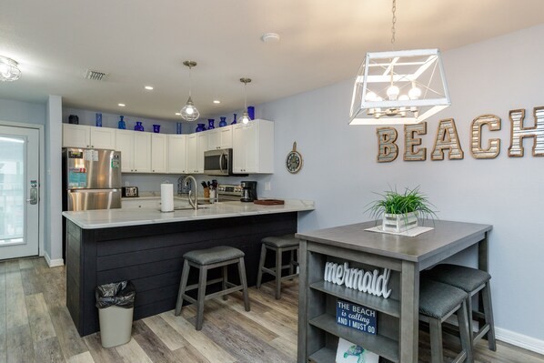 A modern kitchen with a central island, white cabinets, stainless steel appliances, and a 'BEACH' sign on the wall.