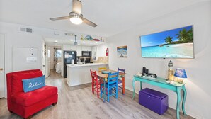 A living room with a red sofa, a wooden dining table with colorful chairs, a teal console table, and a large beach-themed painting on the wall. - Poolside Retreat | King Bed, Private Patio & Relaxation (Corpus Christi)