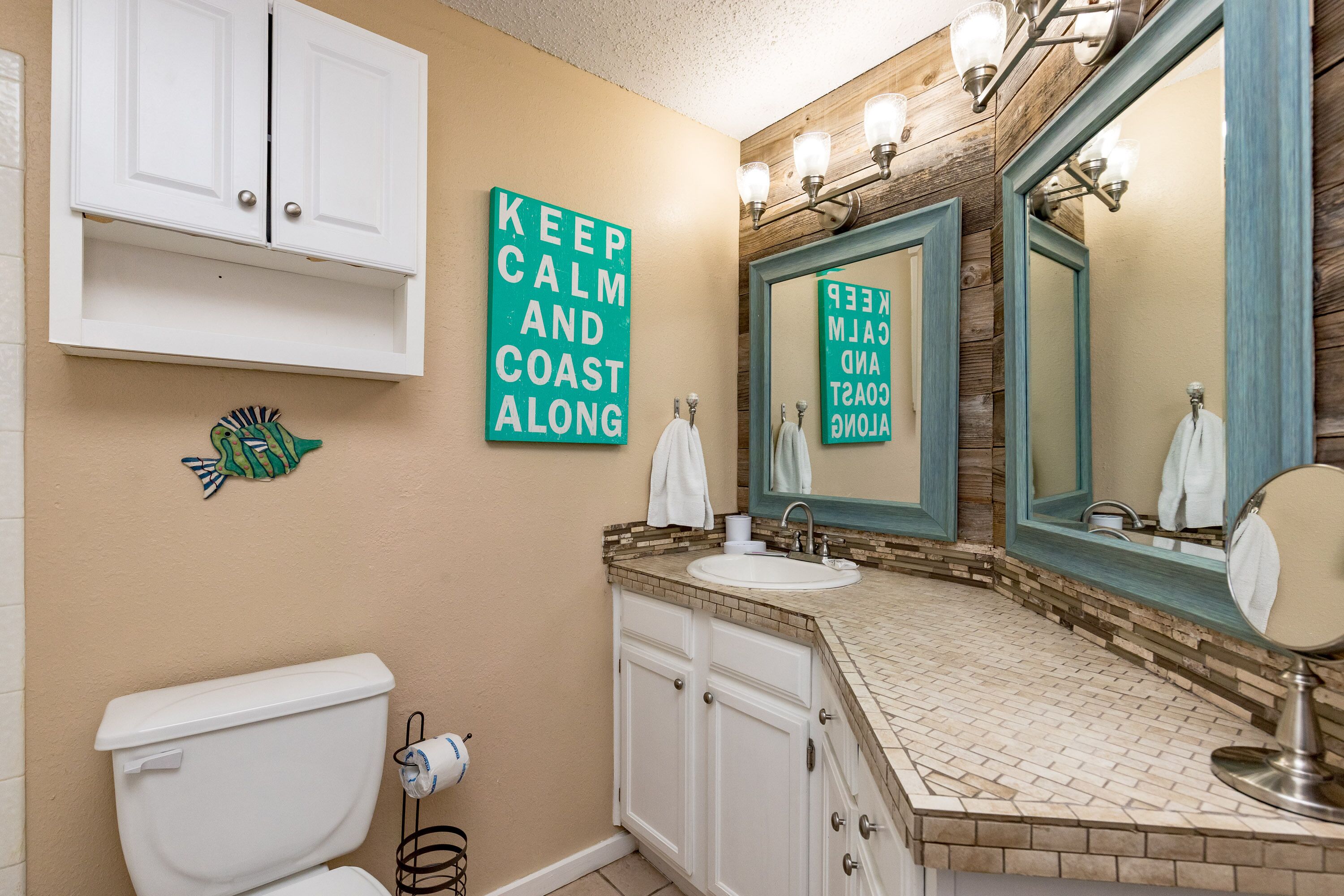 A bathroom with a toilet, sink, and mirror. There are two signs on the wall, one reads 'KEEP CALM AND COAST ALOON' and the other 'KEEP CALM AND COAST ALOON' reflected in the mirror.