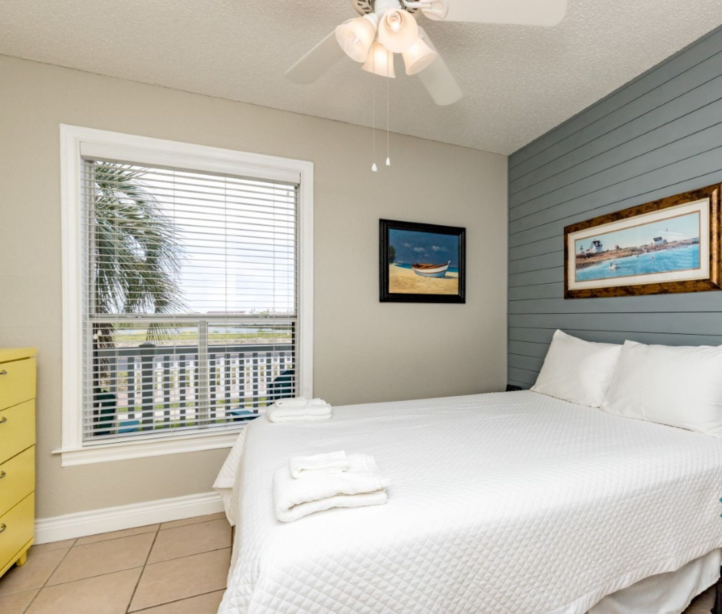 A clean, well-lit bedroom with a bed, a window with blinds, a framed picture on the wall, and a yellow dresser.