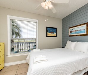 A clean, well-lit bedroom with a bed, a window with blinds, a framed picture on the wall, and a yellow dresser.