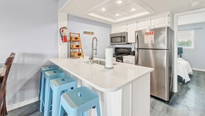 A modern kitchen with white cabinets, a stainless steel refrigerator, a microwave, and a sink. There are blue bar stools and a fire extinguisher on the wall. - End Unit Beach Condo | Pool, Hot Tub & Scenic Dunes Views (Corpus Christi)