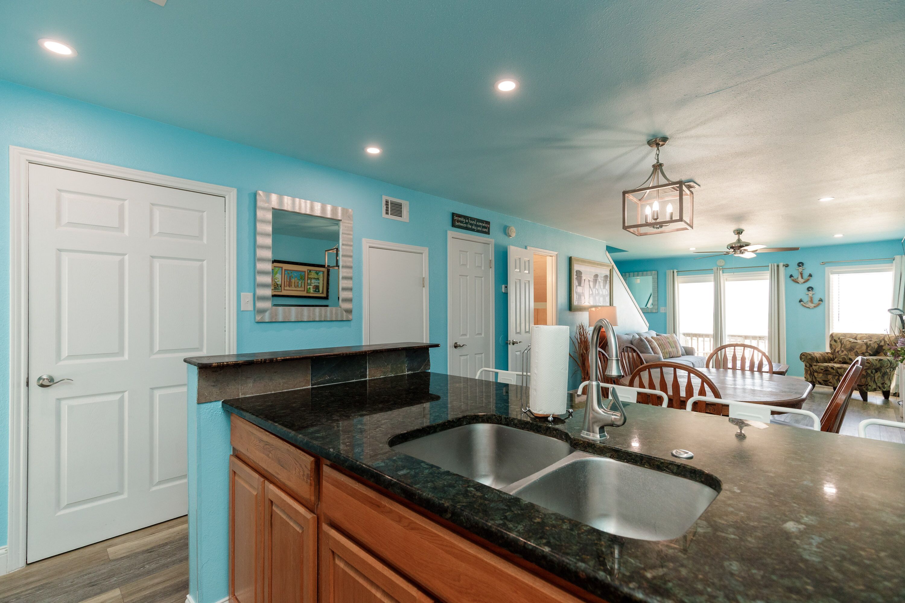 A kitchen with a granite countertop, stainless steel sink, and wooden cabinets. There is a dining area with a table and chairs, and a ceiling fan.
