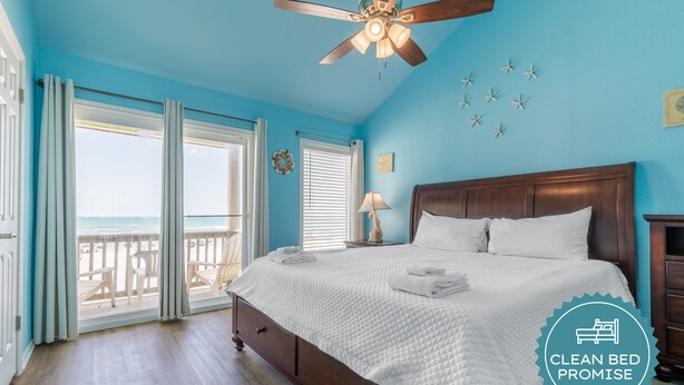 A bedroom with a bed, a ceiling fan, a view of the beach, and a "Clean Bed Promise" badge. - Beachfront Escape | Heated Pool & Gulf Views (Corpus Christi)