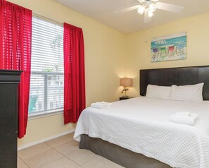 A hotel room with a bed, a nightstand, a lamp, a ceiling fan, and a window with red curtains.
