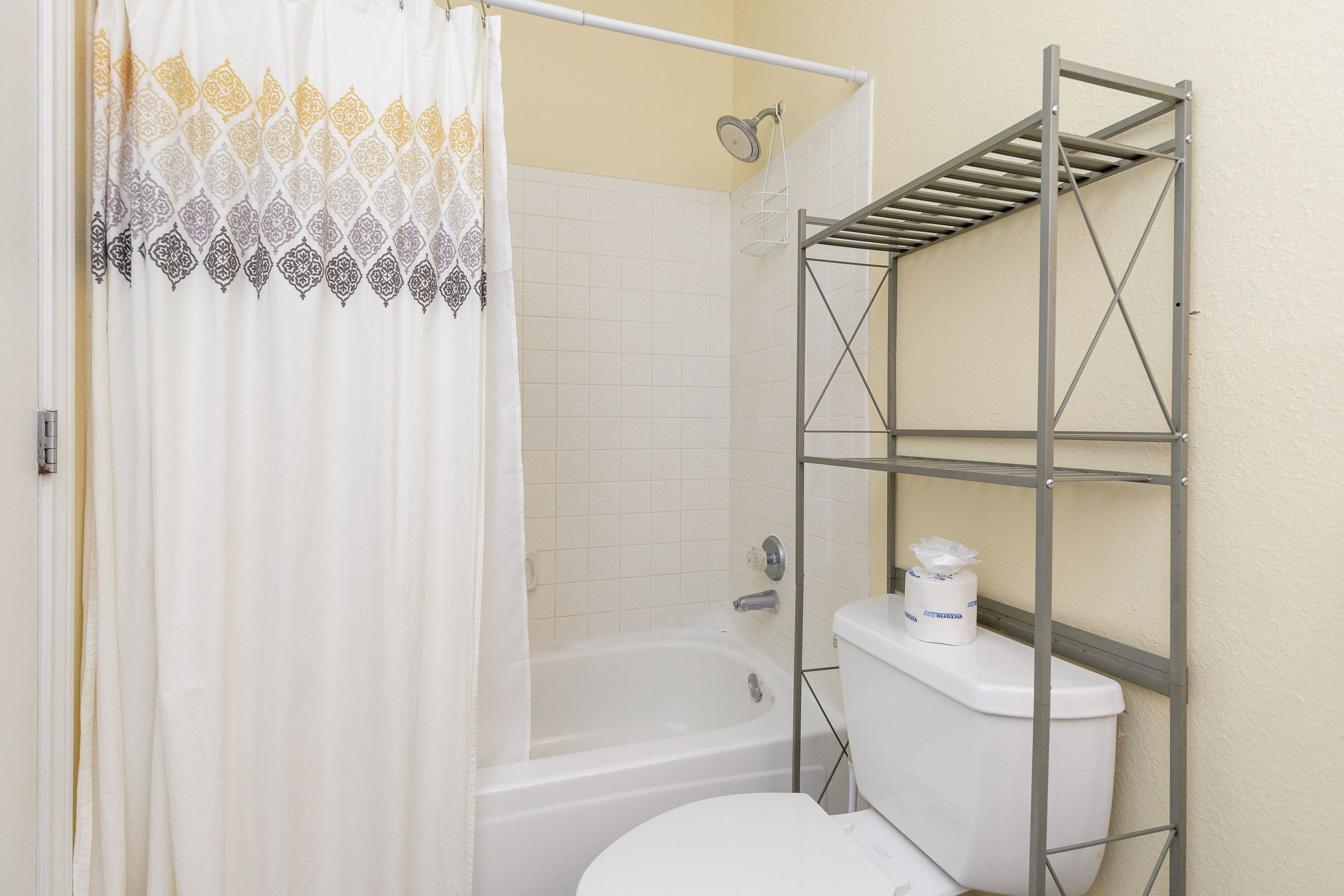 A bathroom with a shower curtain, a bathtub, a toilet, and a metal shelf unit.