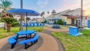 Un espace pique-nique avec des tables et des parasols bleus, entouré de palmiers et d’un bâtiment avec un toit rouge.