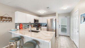 A kitchen with white cabinets, a large island with bar stools, stainless steel appliances, and a 'Beach' sign on the wall. - Beachside Family Escape | Balconies & Steps to Sand (Corpus Christi)