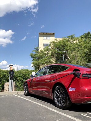 Un véhicule électrique rouge est en cours de recharge à une borne, avec un ciel dégagé et un bâtiment en arrière-plan.