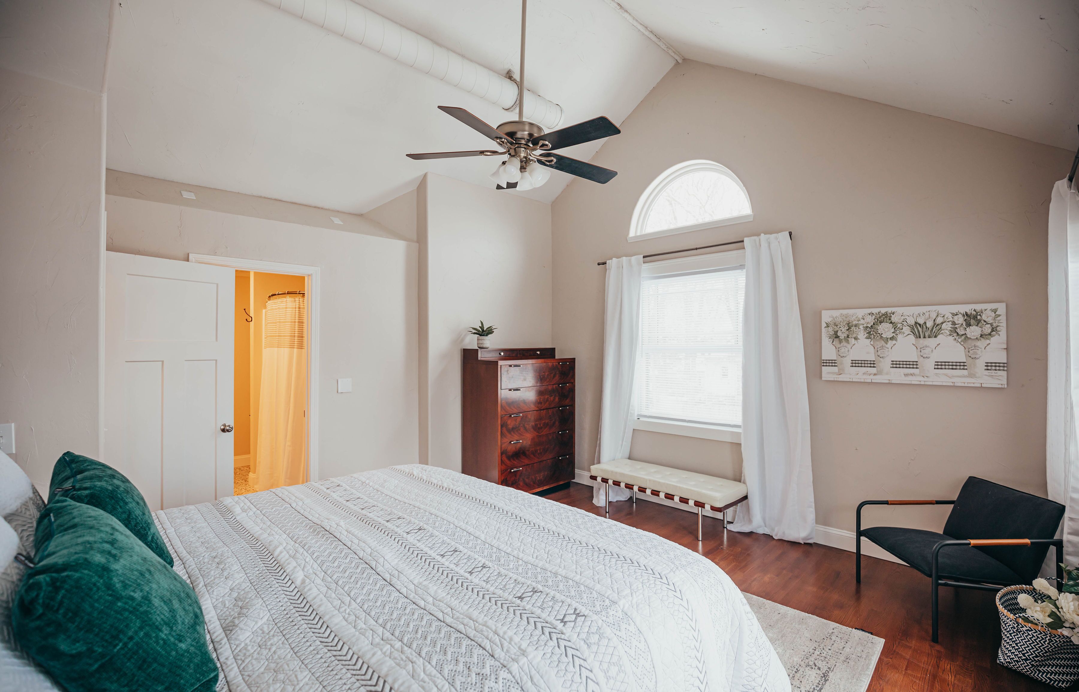 A bedroom with a bed, a dresser, a bench, a chair, a ceiling fan, and a window with curtains.
