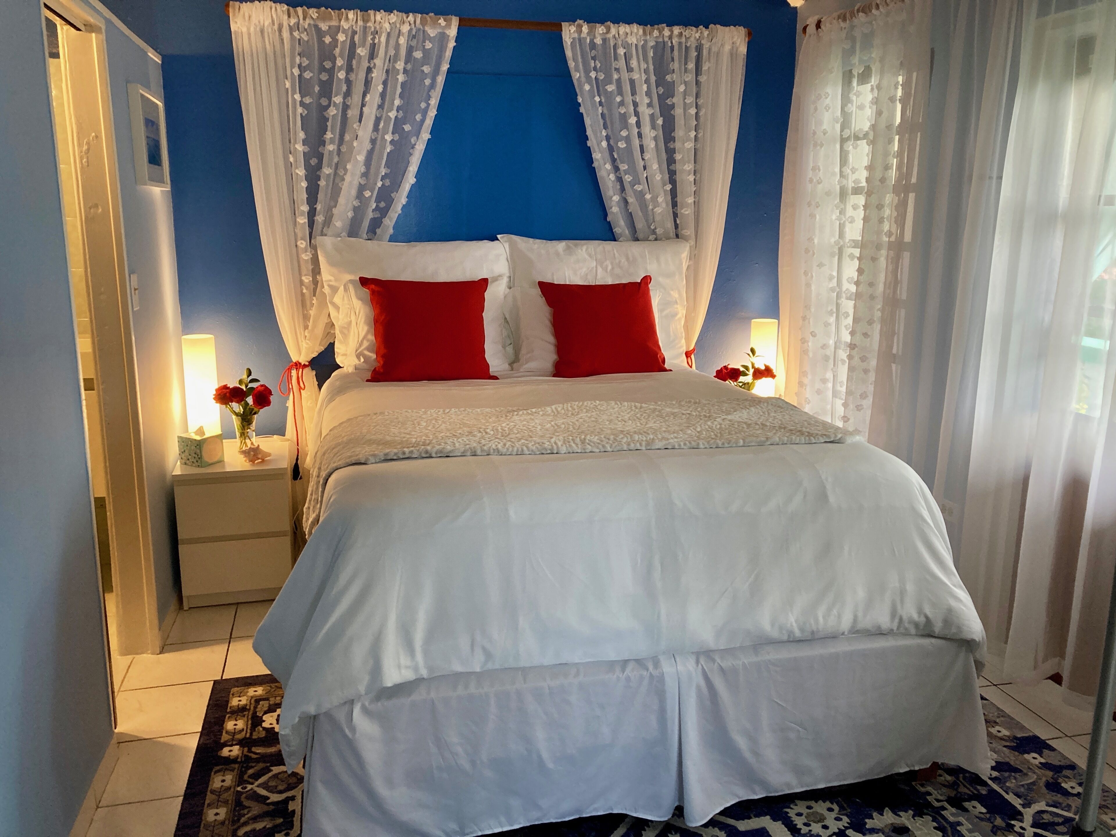 Una cama bien hecha con ropa de cama blanca y roja, una mesita de noche con una lámpara y una ventana con cortinas traslúcidas.