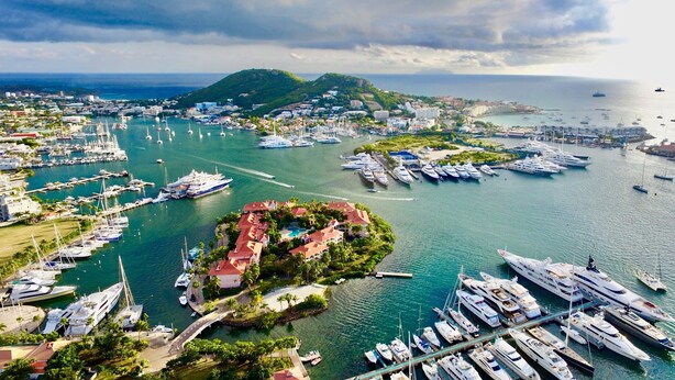 A marina with numerous yachts docked, a small island with buildings, and a view of the coastline.
