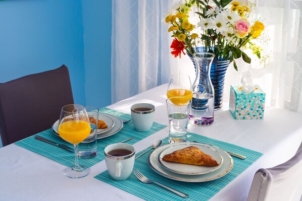 Une table de petit-déjeuner avec un croissant, du jus d’orange, du café et du thé. On y voit un vase rempli de fleurs et une boîte cadeau.