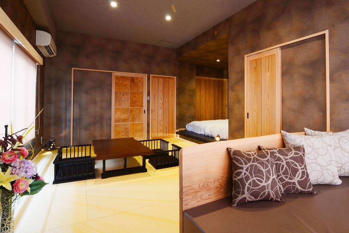 Ein Zimmer mit einem Bett, einem Schlafsofa, einem Tisch und einem Stuhl. Es gibt einen Holzschrank und einen kleinen Tisch mit Blumen in einer Vase.