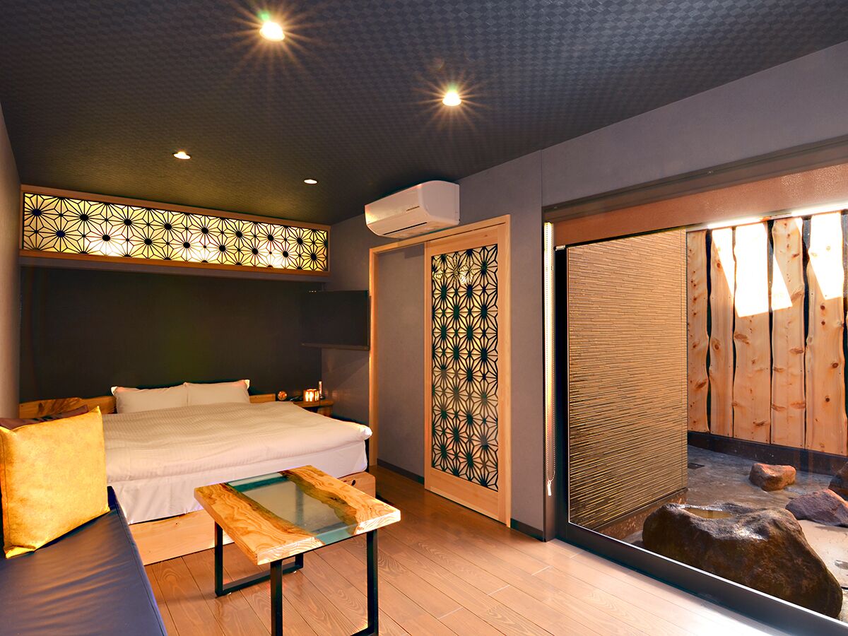 A hotel room with a bed, a small table, a decorative wall panel, and a sliding door leading to a wooden deck.