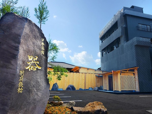 A building with a modern design, featuring a large stone with inscriptions, a wooden structure, and a parking area. - Nihon Ryokan Utsuwa Beppu Kannawa (Beppu)