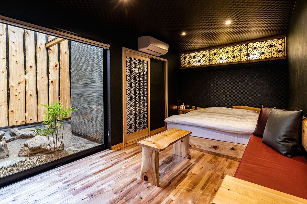 A modern bedroom with a large bed, a wooden bench, and a view of a wooden structure outside.