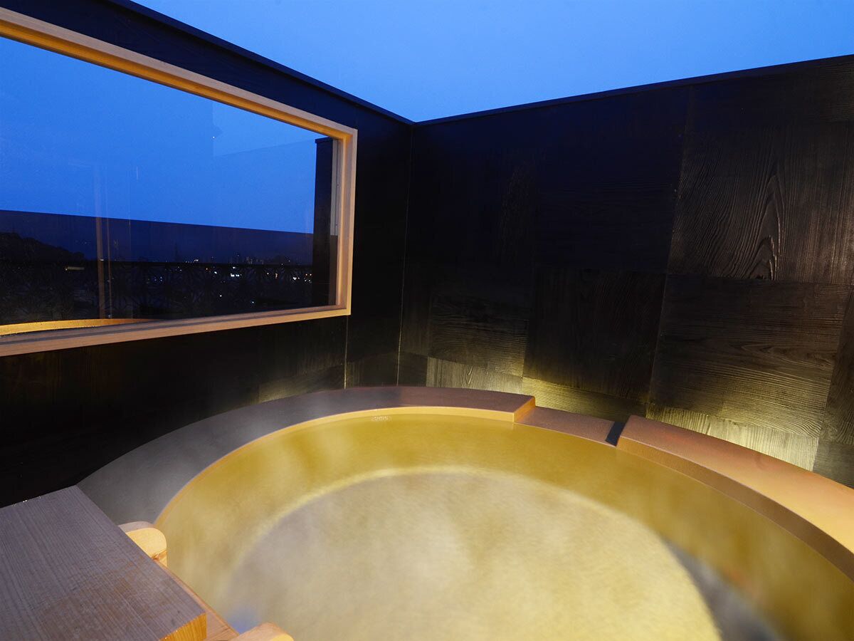 A hot tub in a room with a window overlooking a cityscape at dusk.