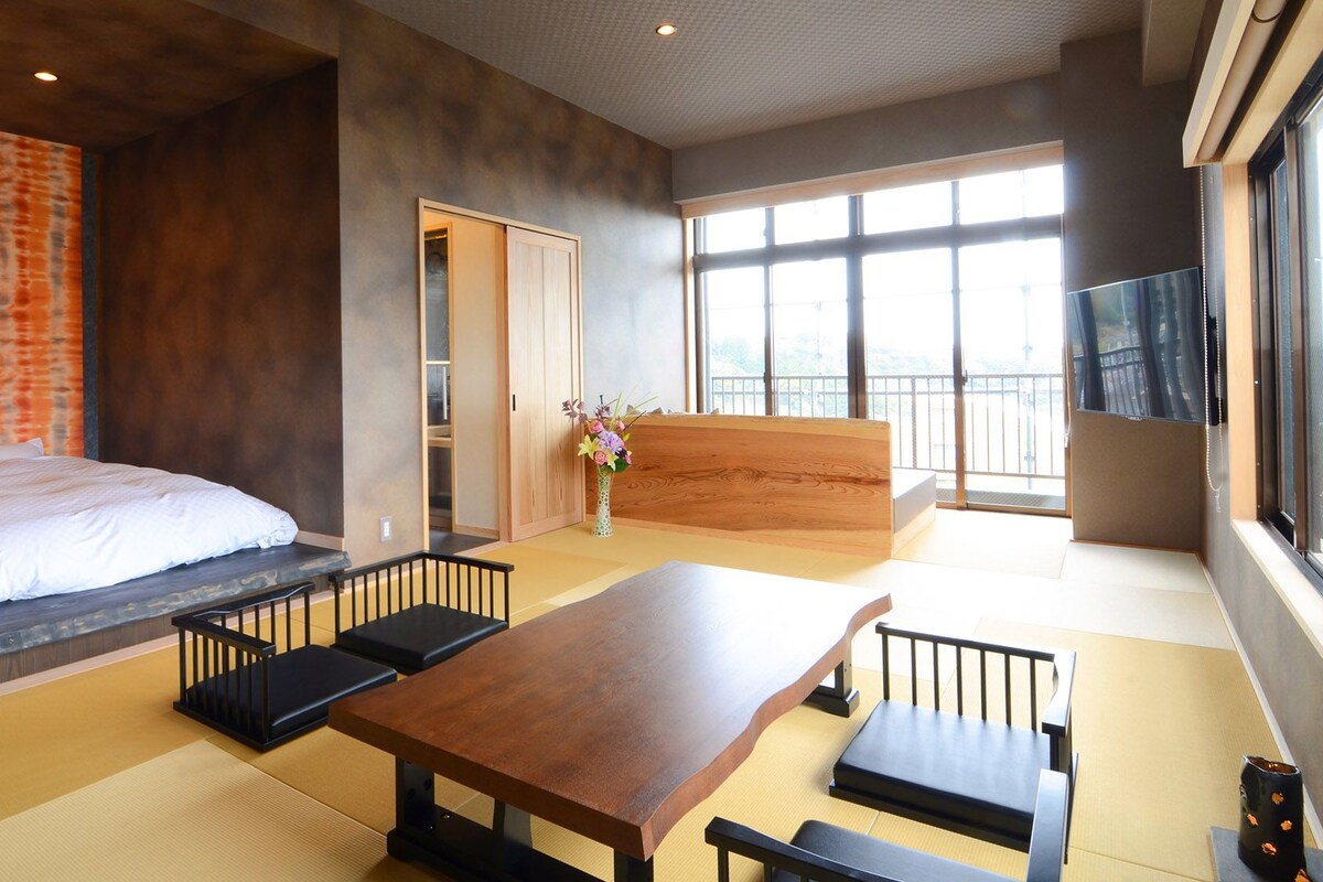 A modern hotel room with a bed, a wooden bench, a table, tatami mats, a sliding door, and a large window.