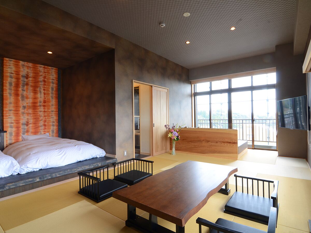 A modern hotel room with a bed, a wooden dining table, tatami flooring, a sliding door, and large windows.