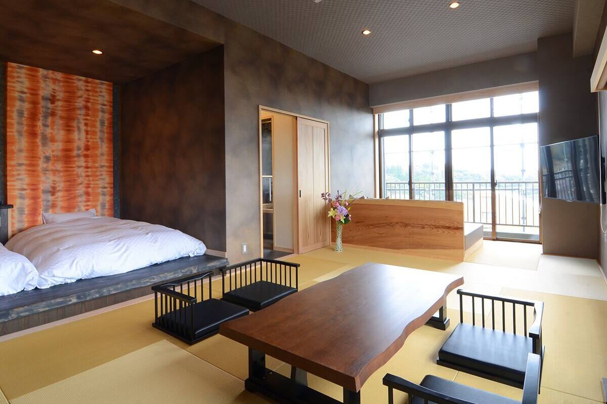 Ein modernes Hotelzimmer mit Bett, Holztisch, Tatami-Boden, einer Schiebetür und großen Fenstern.