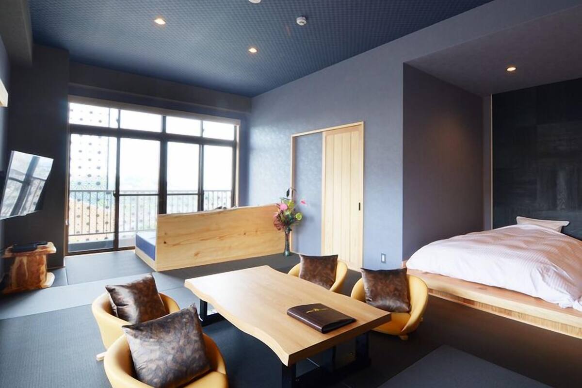 Ein modernes Hotelzimmer mit einem großen Bett, einem Holztisch, zwei Stühlen, einem kleinen Tisch, einem Fenster mit Aussicht und einer Tür.