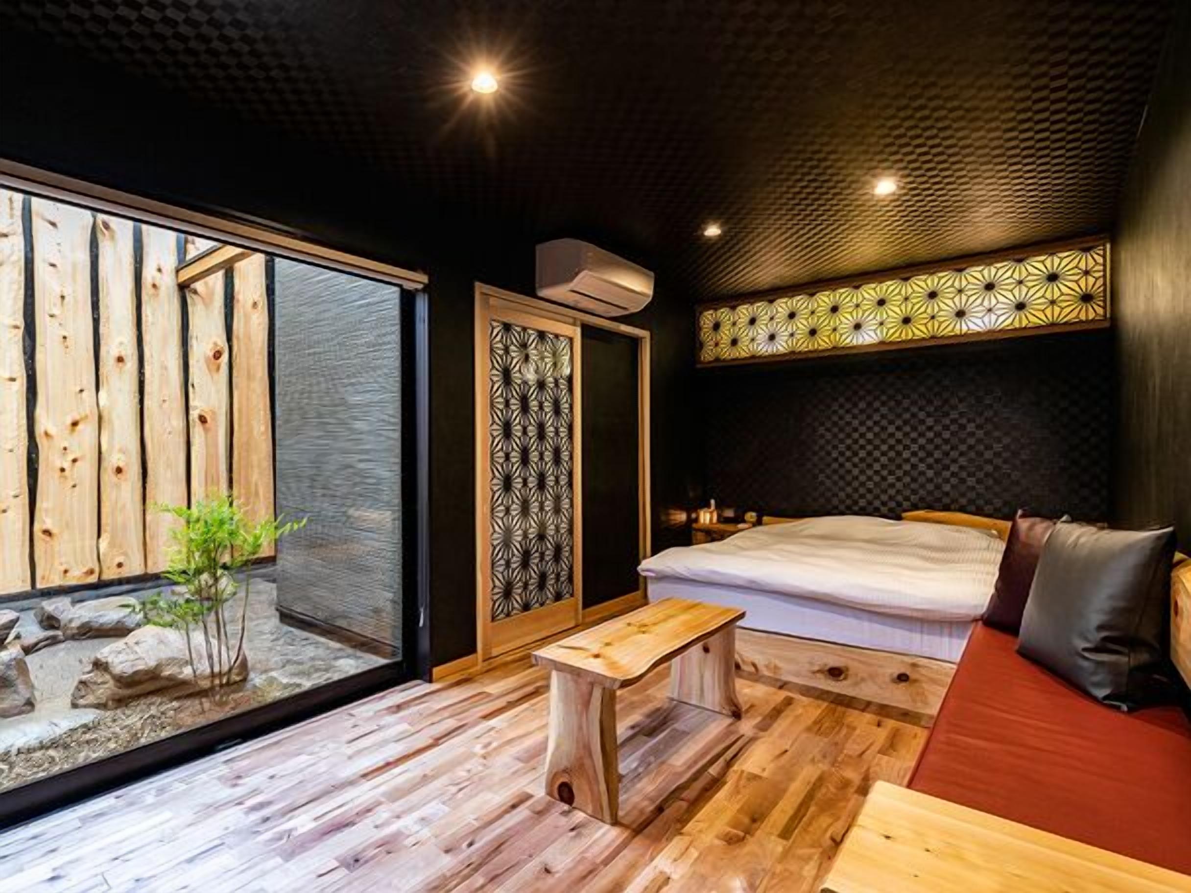 A room with a bed, a wooden bench, a small table, a sliding door, and a view of a wooden structure outside.
