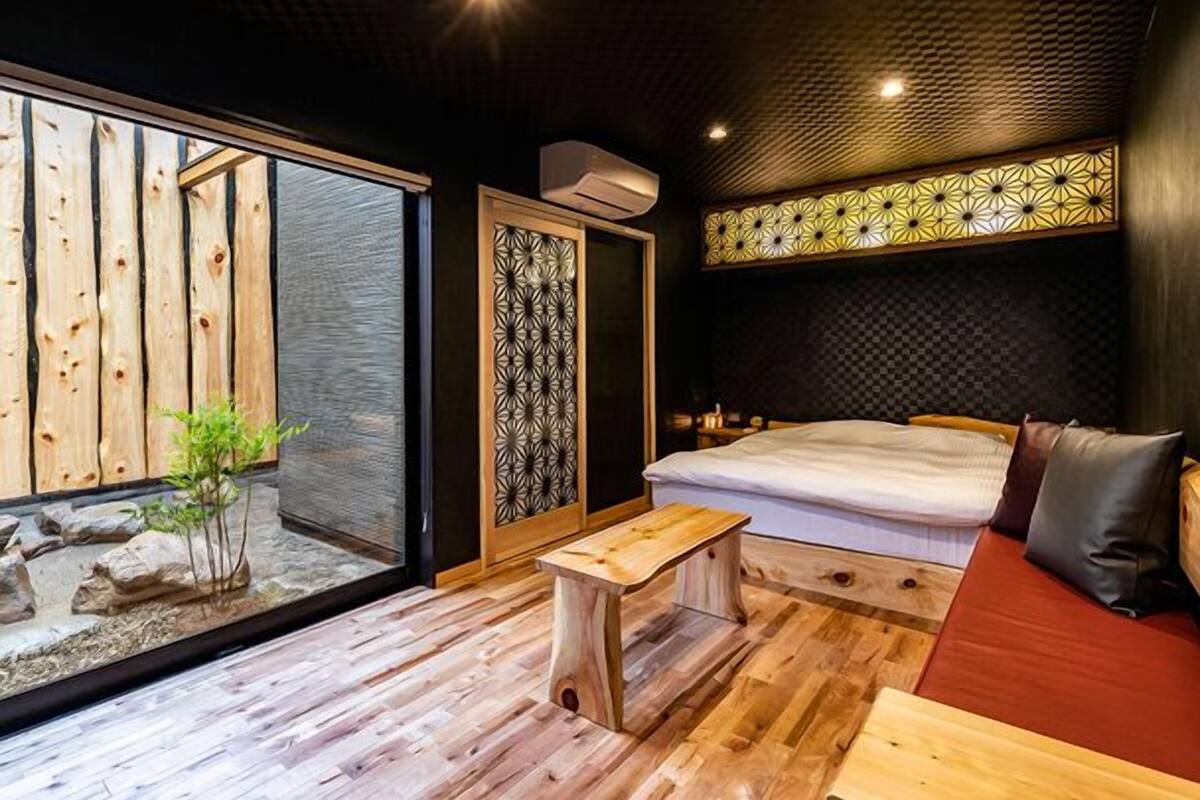 Ein Zimmer mit einem Bett, einer Holzbank, einem kleinen Tisch, einer Schiebetür und Blick auf eine Holzkonstruktion im Freien.