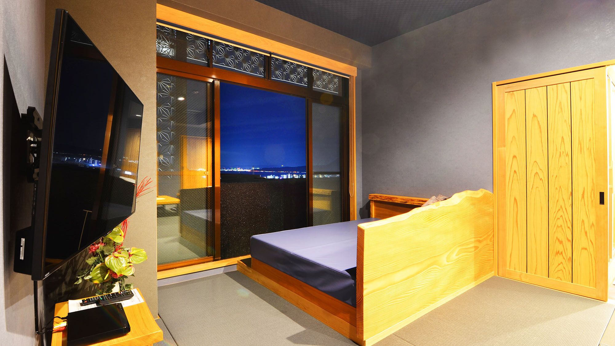 Une chambre à coucher avec un lit, une télévision, un bureau avec un clavier, une plante et une vue sur la ville la nuit.