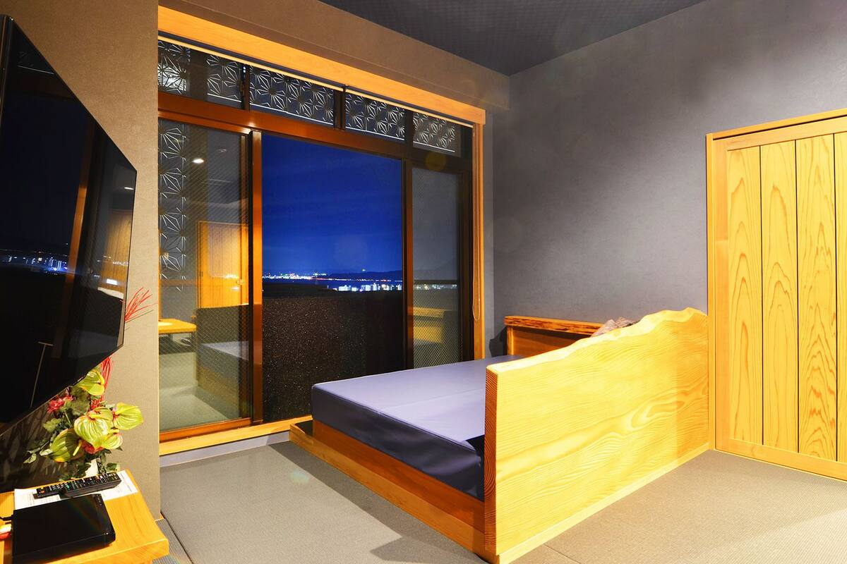 Ein Schlafzimmer mit einem Bett, einem Fernseher, einem Schreibtisch mit Tastatur, einer Pflanze und Blick auf die nächtliche Stadt.