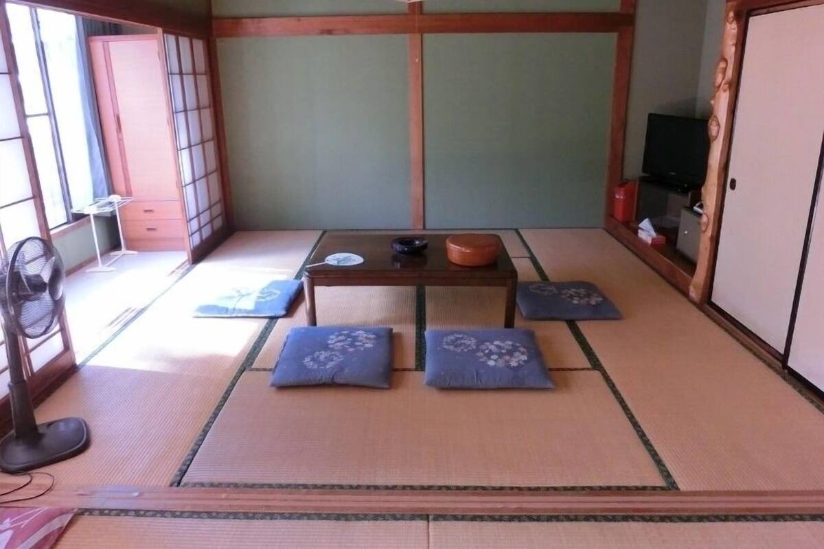 A traditional Japanese room with tatami flooring, a low table, cushions, a fan, and a television.