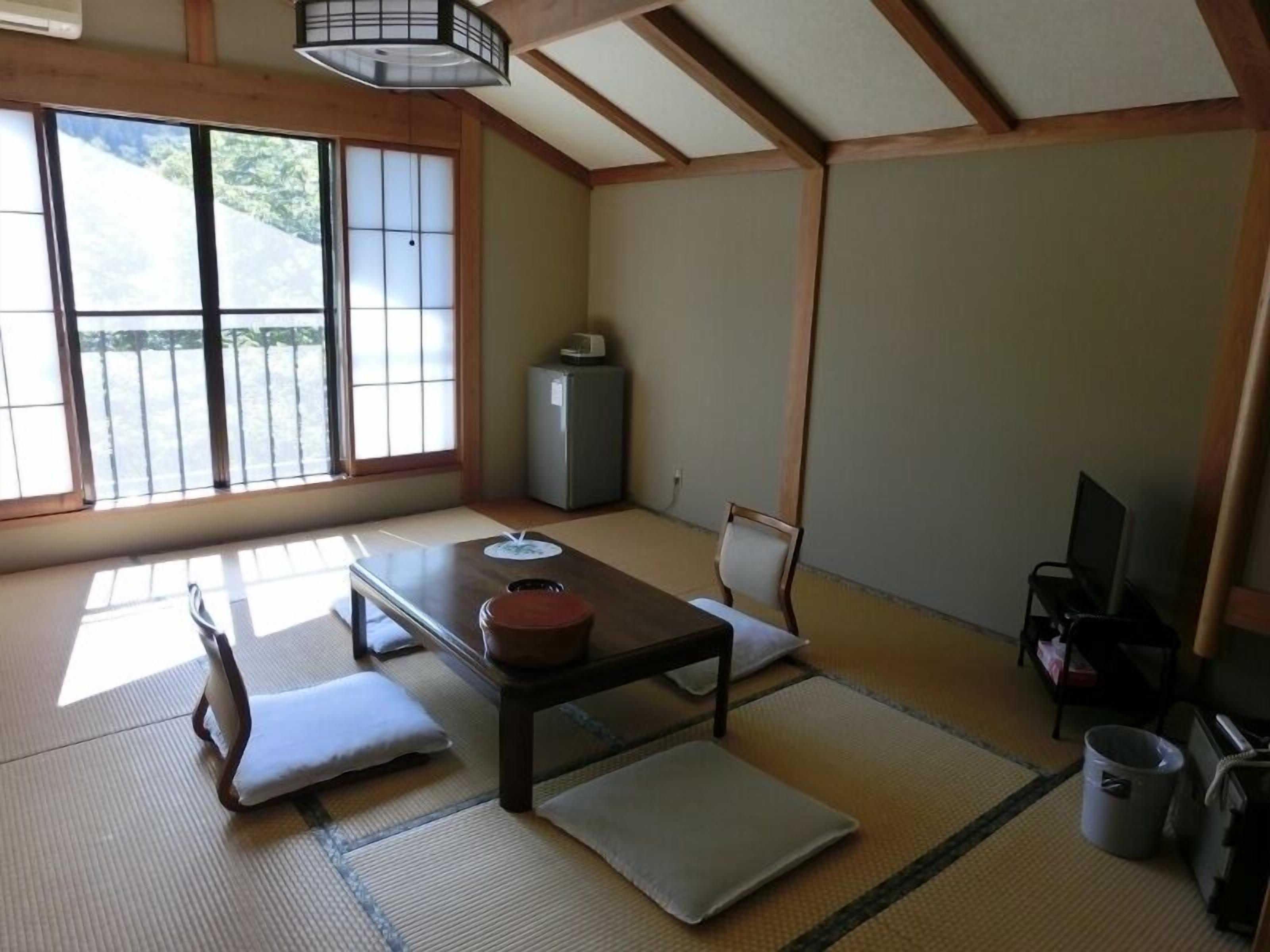 Una habitación tradicional japonesa con suelo de tatami, una mesa baja, un sofá, un televisor y una ventana con vistas a la vegetación.
