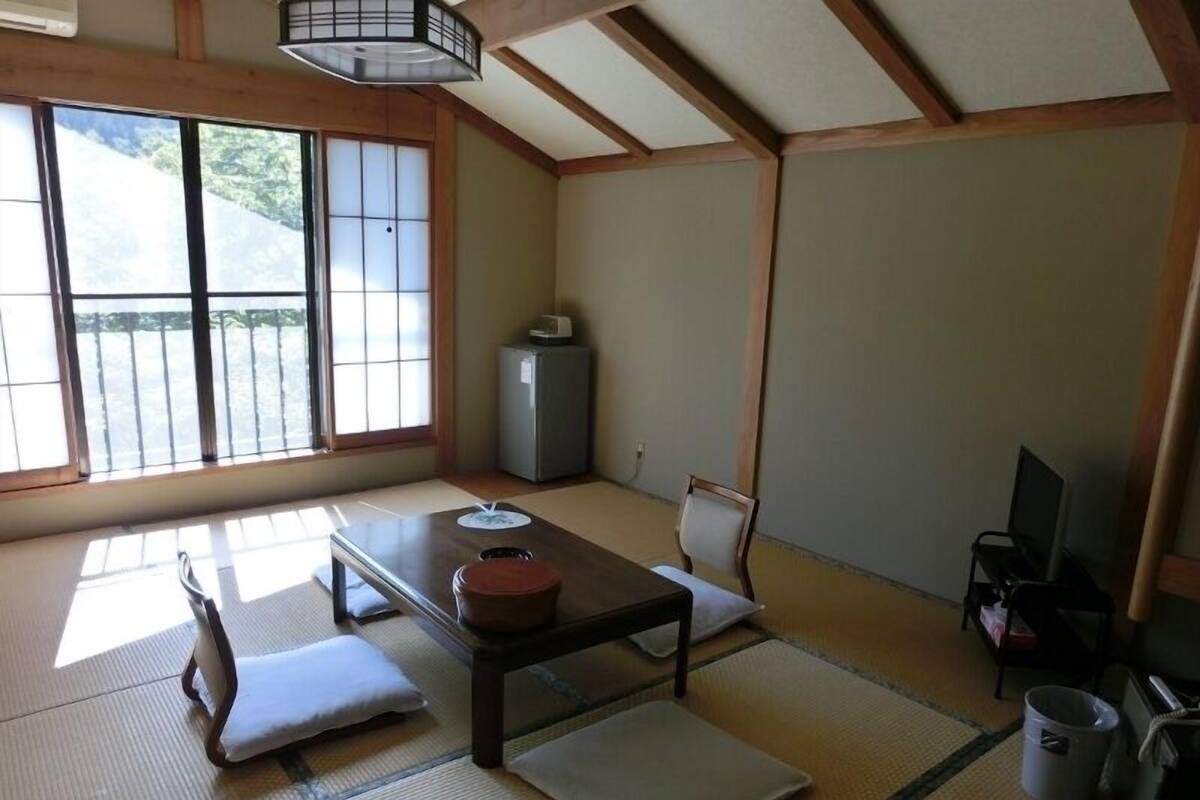 A traditional Japanese room with tatami flooring, a low table, a sofa, a television, and a window with a view of greenery.