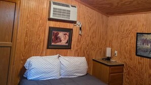 A room with wooden paneling, a bed with a dark quilt, a bedside table with a lamp, and a framed picture on the wall. - Mini Cowboy Cabin (Belfield)