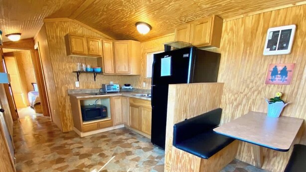 A compact kitchen with wooden cabinets, a black refrigerator, a small dining table, and a stone-tiled floor. - Mini Cowboy Cabin (Belfield)