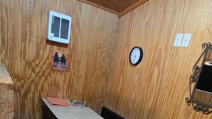 A small room with wooden walls, a stone-tiled floor, a bed with a wooden headboard, a bedside table, a wall-mounted clock, and a wall-mounted air conditioning unit. - Mini Cowboy Cabin (Belfield)