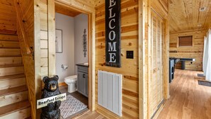 Interior de una cabaña de madera con una escalera, una escultura de oso, un letrero de 'Welcome', un baño y un comedor.