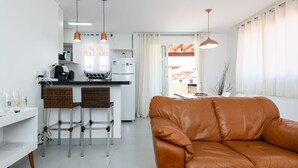 Cocina moderna equipada con refrigerador, cafetera y taburetes de bar. Hay un sofá de cuero marrón y una mesa de comedor con sillas.