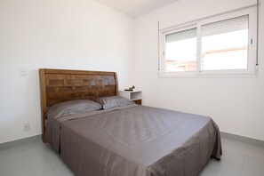 A bedroom with a wooden headboard, a single bed with a grey bedspread, a bedside table, and a window with blinds.