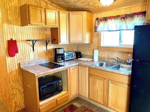 A compact kitchen with wooden cabinets, a microwave, a toaster, a sink, and a refrigerator. - Mini Hunter Cabin (Belfield)