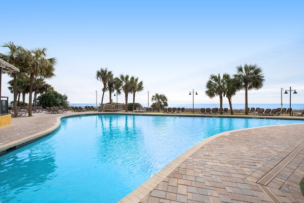 A swimming pool with a curved edge, surrounded by lounge chairs and palm trees. - 17th-floor oceanfront condo with pool, hot tub, WiFi & beach location (Myrtle Beach)