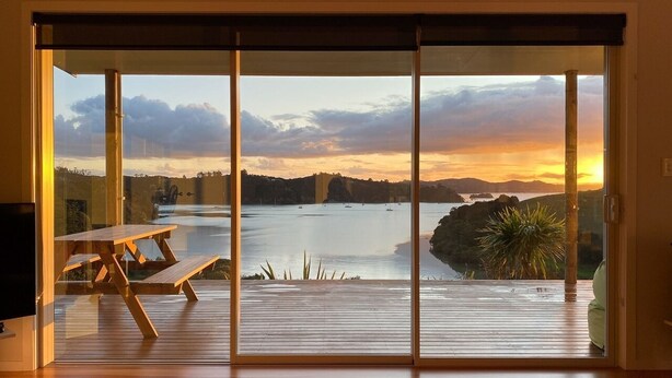 A room with a large sliding glass door leading to a deck overlooking a body of water during sunset. - Magic Water Views Russell (Russell)