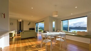 A modern living room with a wooden floor, a green sofa, a dining table with chairs, and large windows offering a view of a lake and hills. - Magic Water Views Russell (Russell)