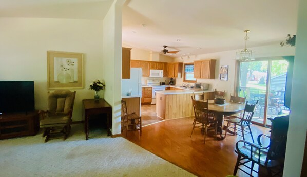 A spacious living room with a large TV, a dining area with wooden chairs, and a kitchen with wooden cabinets and a central island.