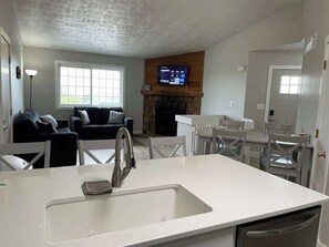 Een moderne keuken met een wit aanrecht, een roestvrijstalen spoelbak en een stenen open haard. Een flatscreen televisie is boven de open haard bevestigd.