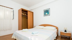 A neatly made bed with a white bedspread, a wooden wardrobe, a bedside table with a lamp, and a framed picture on the wall. - Guesthouse Home Sweet Home - Standard One-Bedroom Apartment with Balcony (Dubrovnik)