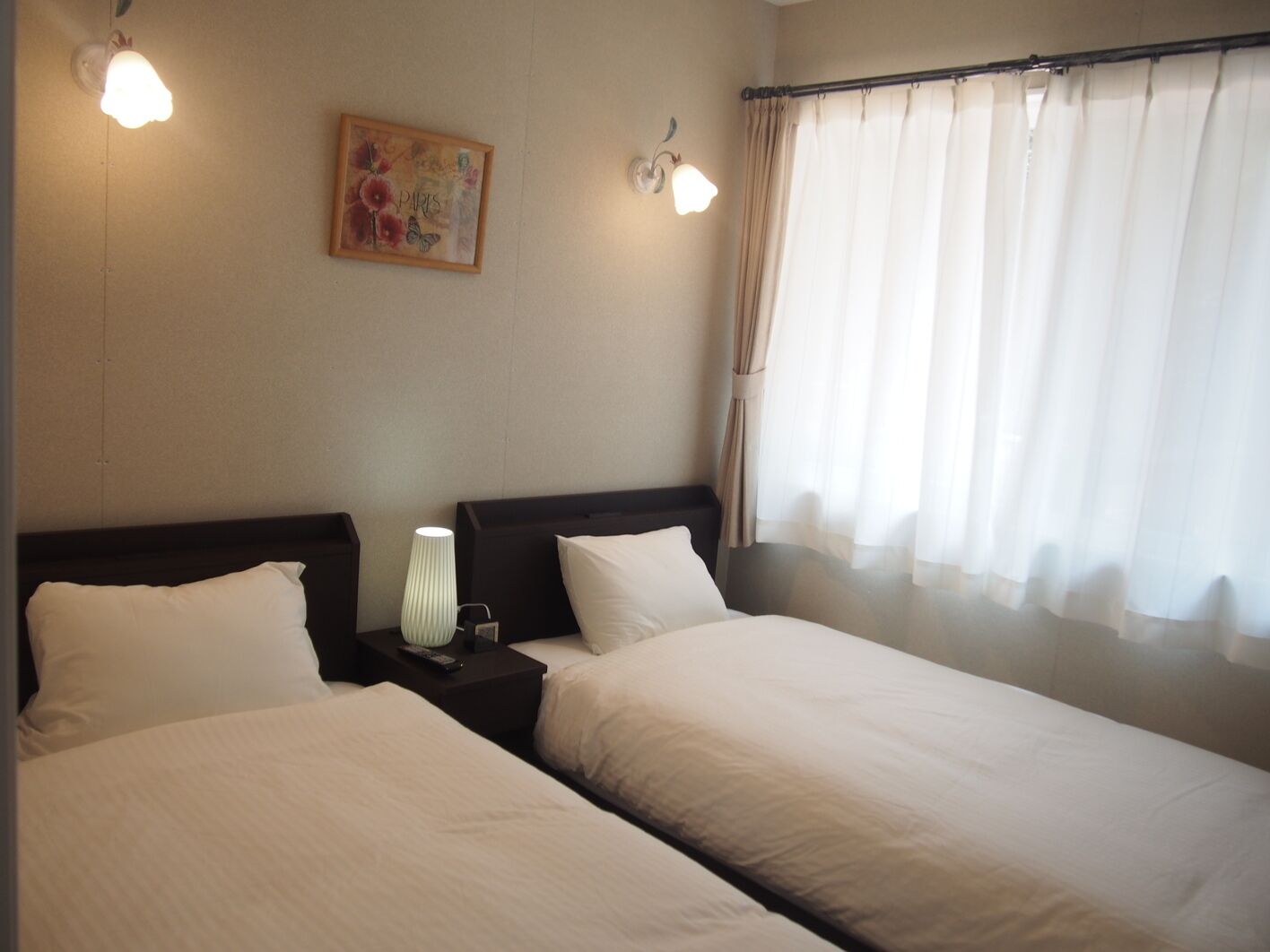 Habitación de hotel con dos camas, una mesita de noche con lámpara, un cuadro en la pared y una ventana con cortinas.