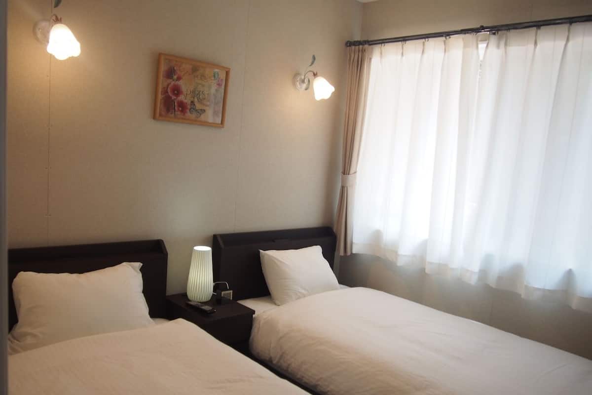 Een hotelkamer met twee bedden, een nachtkastje met een lamp, een schilderij aan de muur en een raam met gordijnen.